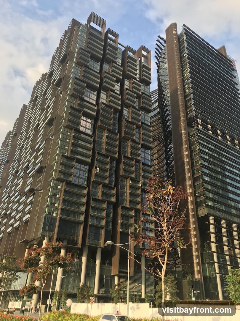 A low-angle shot of two modern high-rise buildings against a cloudy sky. The building on the left has a distinctive facade with numerous balconies and a bronze-colored exterior. The building on the right is taller and has a sleek, glass-covered exterior. Trees and streetlights are visible at the base of the buildings.