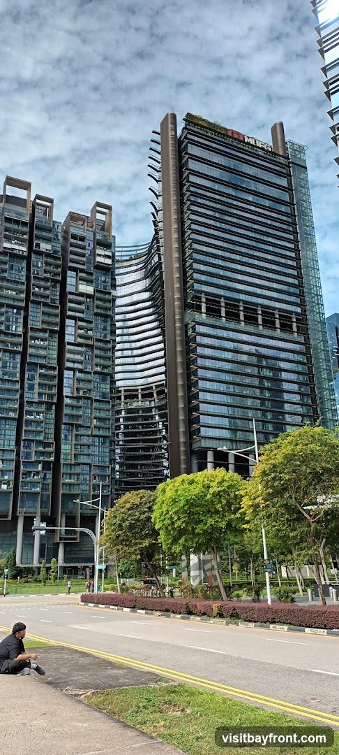 A view of a modern cityscape featuring several tall buildings. The prominent building on the right has the MUFG logo visible at the top. The buildings have a glass facade and a contemporary architectural design. In the foreground, a person is sitting on the sidewalk next to a road, with trees and landscaping lining the street. The sky is partly cloudy.