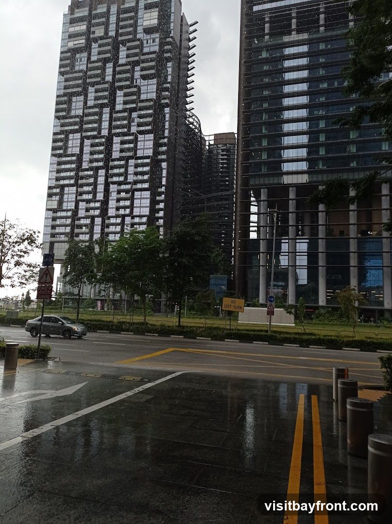 A view of a city street on a rainy day, with modern high-rise buildings in the background. The wet pavement reflects the overcast sky and the surrounding structures. A car drives on the road, and traffic signs are visible. Trees line the street, adding a touch of greenery amidst the urban landscape.