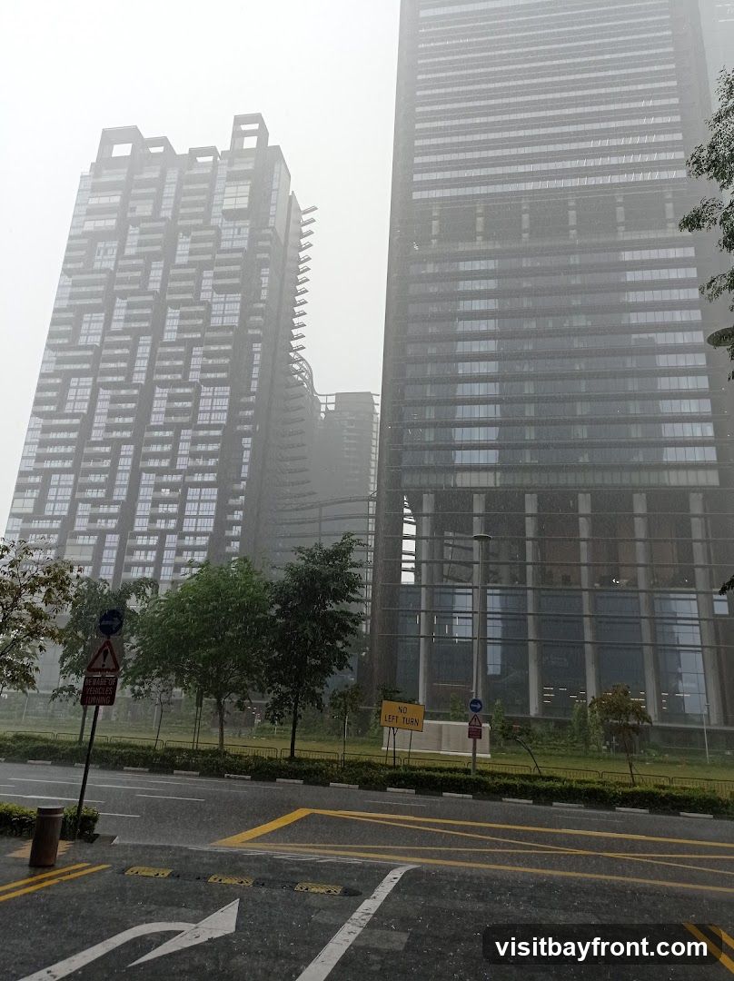 A view of modern skyscrapers on a foggy and rainy day. The rain is visible as streaks against the buildings and on the wet road in the foreground. Traffic signs indicating "BEWARE OF VEHICLES TURNING" and "NO LEFT TURN" are present.