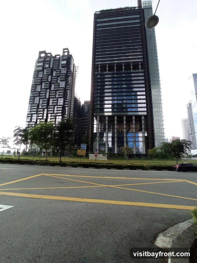 A view of several modern skyscrapers with distinctive architectural designs. The buildings are tall and feature glass facades. In the foreground, there is a road with yellow markings and a black car driving by. The sky is overcast.