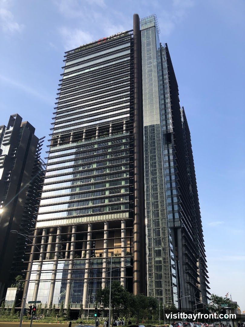 A low-angle shot of a tall, modern skyscraper with a glass facade and horizontal louvers. The building has a sleek, contemporary design, with a prominent glass elevator shaft running vertically along one side. The sky is clear and blue, with a few wispy clouds. The base of the building features some greenery and a street with traffic and pedestrians.
