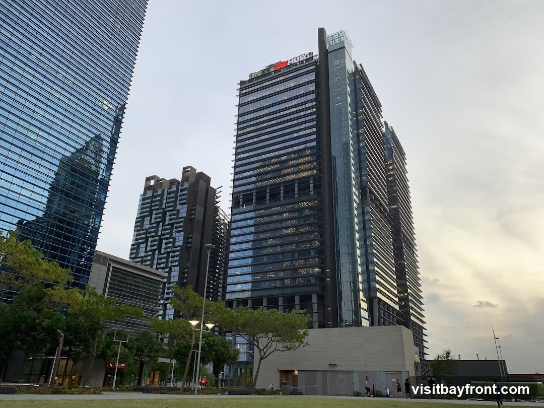 A view of several modern skyscrapers with glass facades, reflecting the sky and surrounding buildings. In the foreground, there is a well-maintained green lawn with scattered trees and lampposts. A few people are visible walking in the distance, suggesting a public space or business district. The sky is overcast with hints of sunlight breaking through. One of the buildings has a visible logo at the top that reads 'MUFG'.