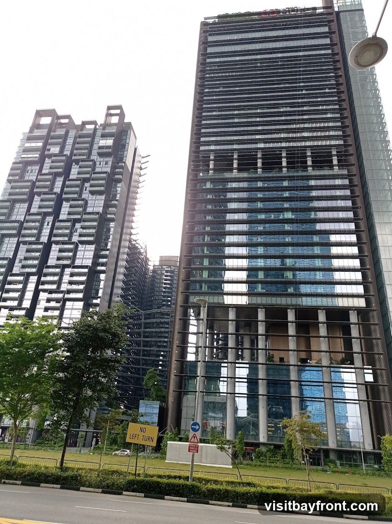 A view of several modern skyscrapers with glass facades, reflecting the sky and surrounding buildings. In the foreground, there is a road with a grassy median strip. A yellow sign with the text "NO LEFT TURN" is visible, along with other traffic signs. Trees and landscaping are present around the base of the buildings.