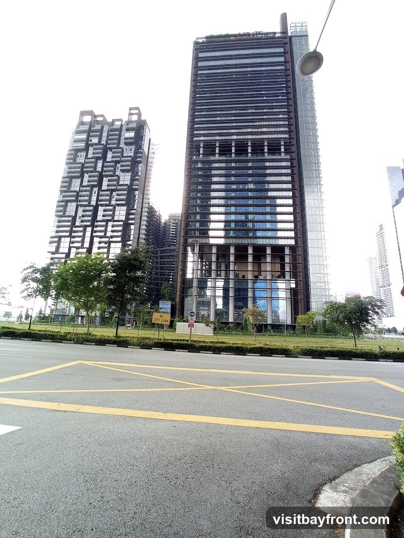 A view of several modern skyscrapers with glass facades and intricate architectural designs. The buildings are tall and imposing, set against a bright, overcast sky. In the foreground, a road with yellow lane markings is visible, along with a green median strip and some trees. A traffic sign indicating "NO LEFT TURN" is also present.