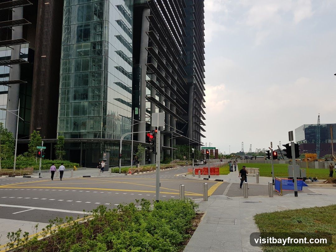 A wide shot of a modern, glass-fronted office building with a distinctive slatted exterior. The building is situated next to a street intersection with traffic lights and pedestrian crossings. In the background, construction cranes and other buildings are visible under a cloudy sky. Several people are walking on the sidewalk and crossing the street. The foreground features green landscaping and a paved walkway.