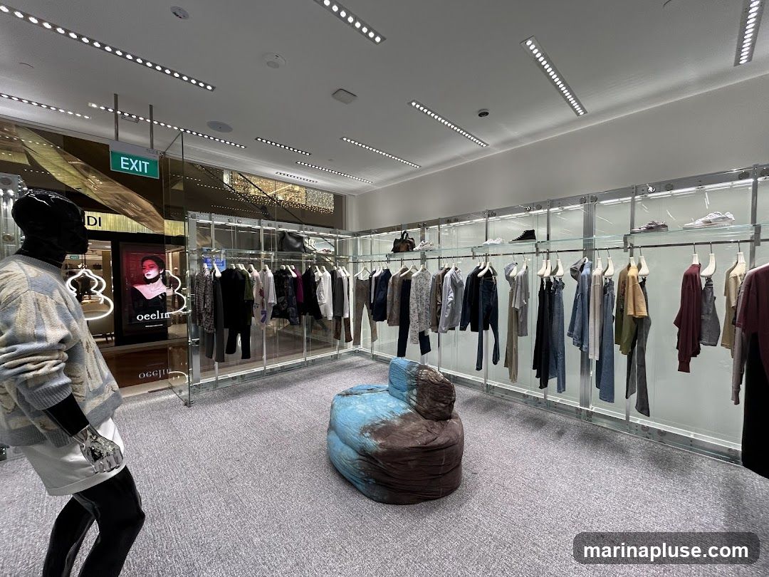 A view inside a modern clothing store, featuring a mannequin dressed in a patterned sweater and white shorts, a display of clothing on racks, and a unique blue and brown beanbag chair. The store has a clean, minimalist design with bright overhead lighting and glass display shelves showcasing shoes. An "EXIT" sign is visible in the background, along with a digital display featuring a woman's portrait and the word "oeelin" illuminated.