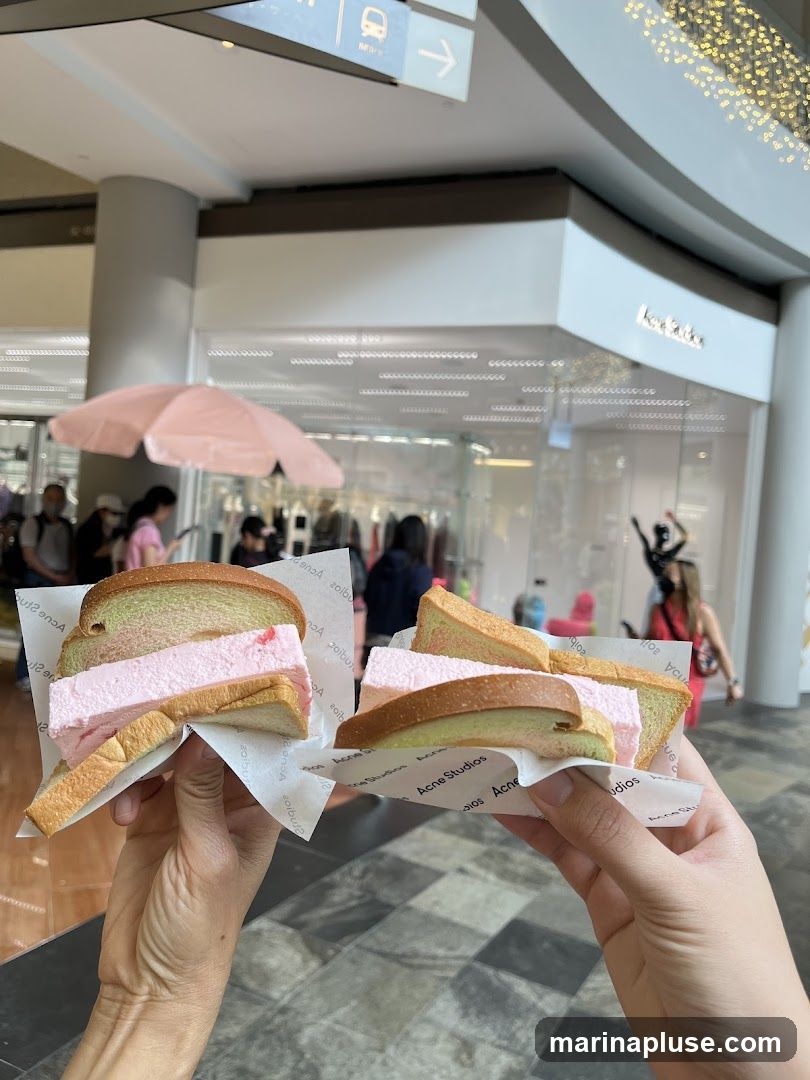 Two ice cream sandwiches are held up in front of a store. The sandwiches are made with pink ice cream between slices of bread. The bread appears to be lightly toasted. The store in the background has a sign that says "Acne Studios".