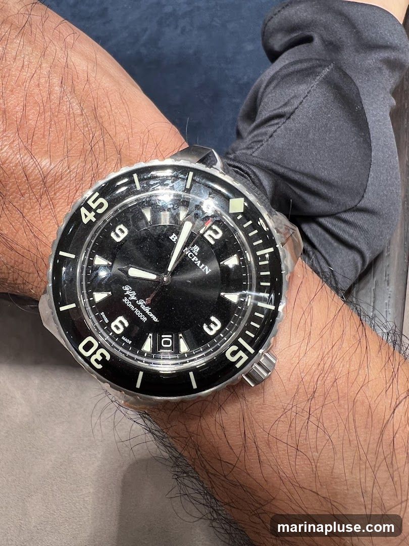 A close-up shot of a black-faced watch with a rotating bezel on a person's wrist. The watch has white hour markers and hands, and the brand name "BLANCPAIN" is visible on the dial, along with "Fifty Fathoms" and "300m/1000ft". The watch is partially covered in clear plastic protective film.