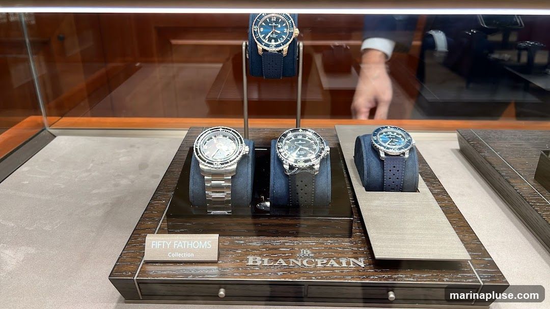 A display case featuring several Blancpain Fifty Fathoms watches. The watches are presented on dark blue velvet stands, with some on a wooden display base. The base has the text "FIFTY FATHOMS Collection" and "BLANCPAIN" visible. A person in a suit is partially visible in the background reflection.