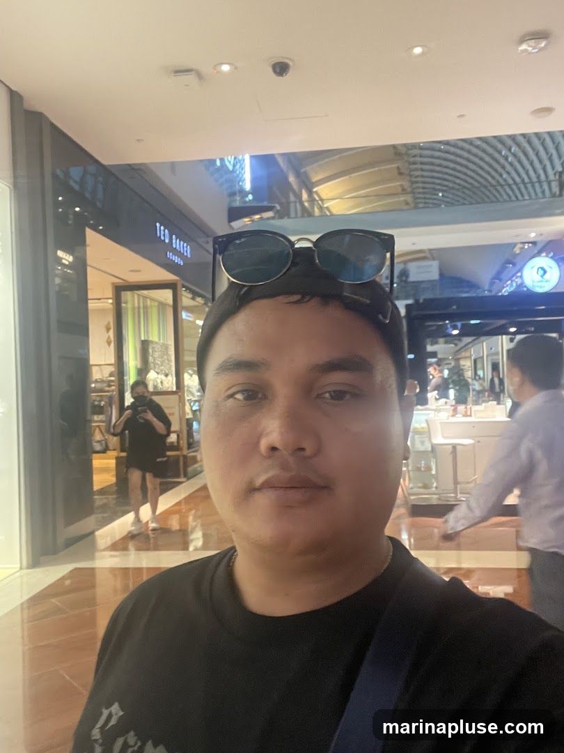 A man takes a selfie in a shopping mall. He is wearing a black t-shirt and has sunglasses on top of his head. In the background, there are various shops, including one with a "TED BAKER LONDON" sign. Other shoppers are visible in the distance.