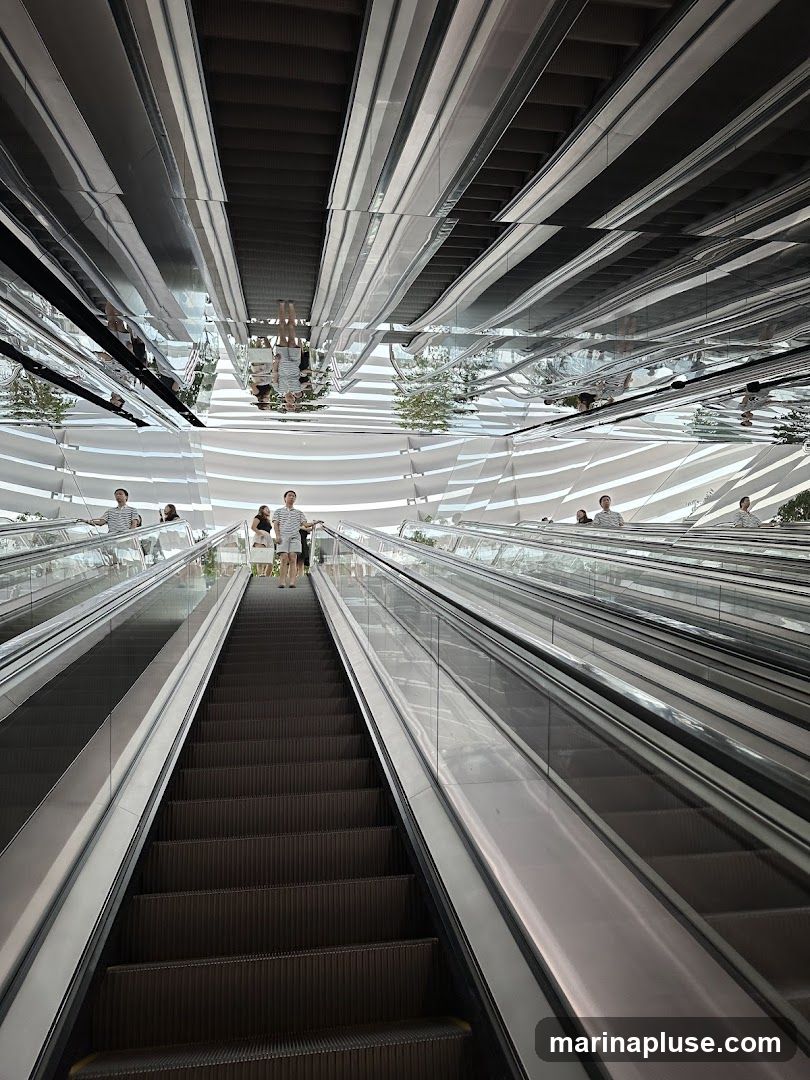 A wide shot of multiple escalators moving upwards, with a highly reflective, mirrored ceiling that creates a disorienting and visually striking effect. People are visible on the escalators, their reflections distorted in the ceiling. The architecture features curved, white walls with horizontal lines.