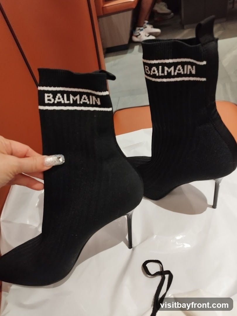 A pair of black Balmain sock boots with white lettering on the ankle cuff. The boots have pointed toes and stiletto heels. They are placed on a white surface, possibly a shopping bag, with a black string in the foreground. In the background, a person is seated on an orange chair.