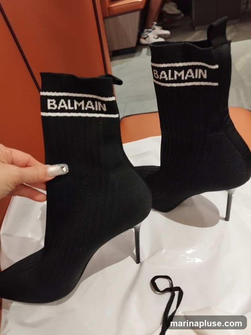 A pair of black Balmain sock boots with white lettering on the ankle cuff. The boots have pointed toes and stiletto heels. They are placed on a white surface, possibly a shopping bag, with a black string in the foreground. In the background, a person is seated on an orange chair.