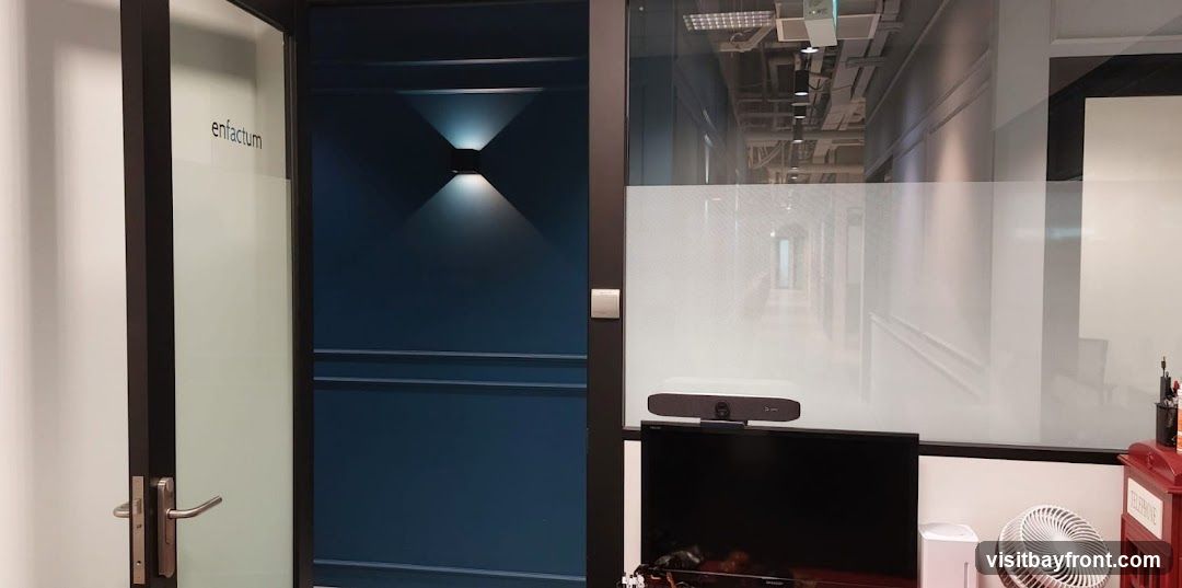 The image shows an office interior with a focus on a doorway and a conference setup. The doorway is dark blue with a modern wall light above it. To the right of the door, a glass partition reveals a hallway with exposed ceiling infrastructure. A flat-screen monitor is visible, topped with a conference bar and camera. Below the monitor, a white air purifier and a white fan are placed on a desk. A red telephone booth-shaped object is also present on the desk. The word "enfactum" is visible on a frosted glass panel near the door.