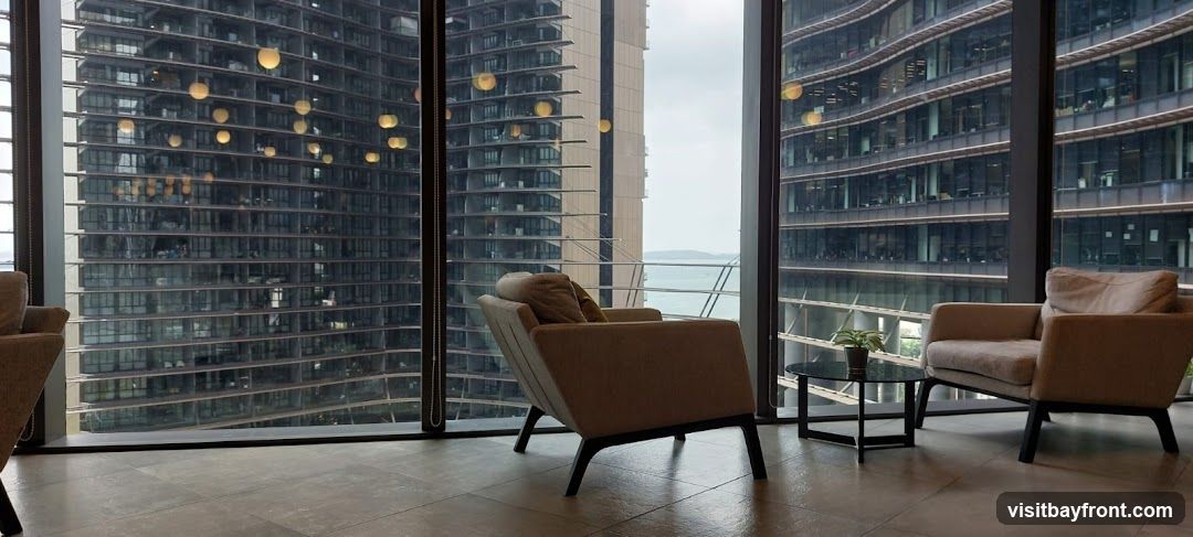 A contemporary lounge area with two comfortable armchairs and a small coffee table, set against a backdrop of modern skyscrapers and a body of water. The interior features large windows offering panoramic views of the city and the sea. Pendant lights are visible in the distance, adding to the ambiance.