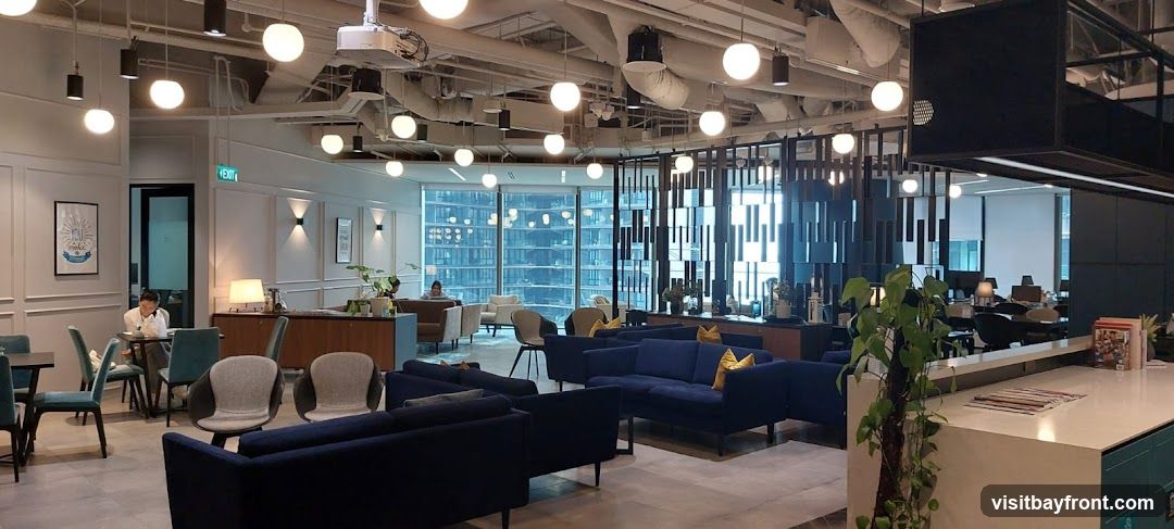 An interior view of a contemporary co-working space, featuring a lounge area with navy blue sofas and yellow accent pillows, comfortable armchairs, and small tables. Large windows offer a view of a city skyline. The space is well-lit with numerous pendant lights and has a modern aesthetic with decorative screens and plants. In the background, an office area with desks and computers is visible.