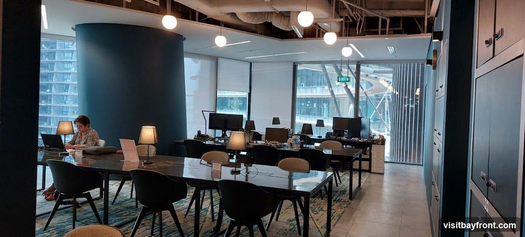 An interior view of a contemporary office space with multiple workstations and a communal work table. A woman is seated at the table working on a laptop, illuminated by a desk lamp. The room features large windows offering views of a city skyline, modern pendant lighting, and dark cabinetry along one wall. An exit sign is visible above the windows.