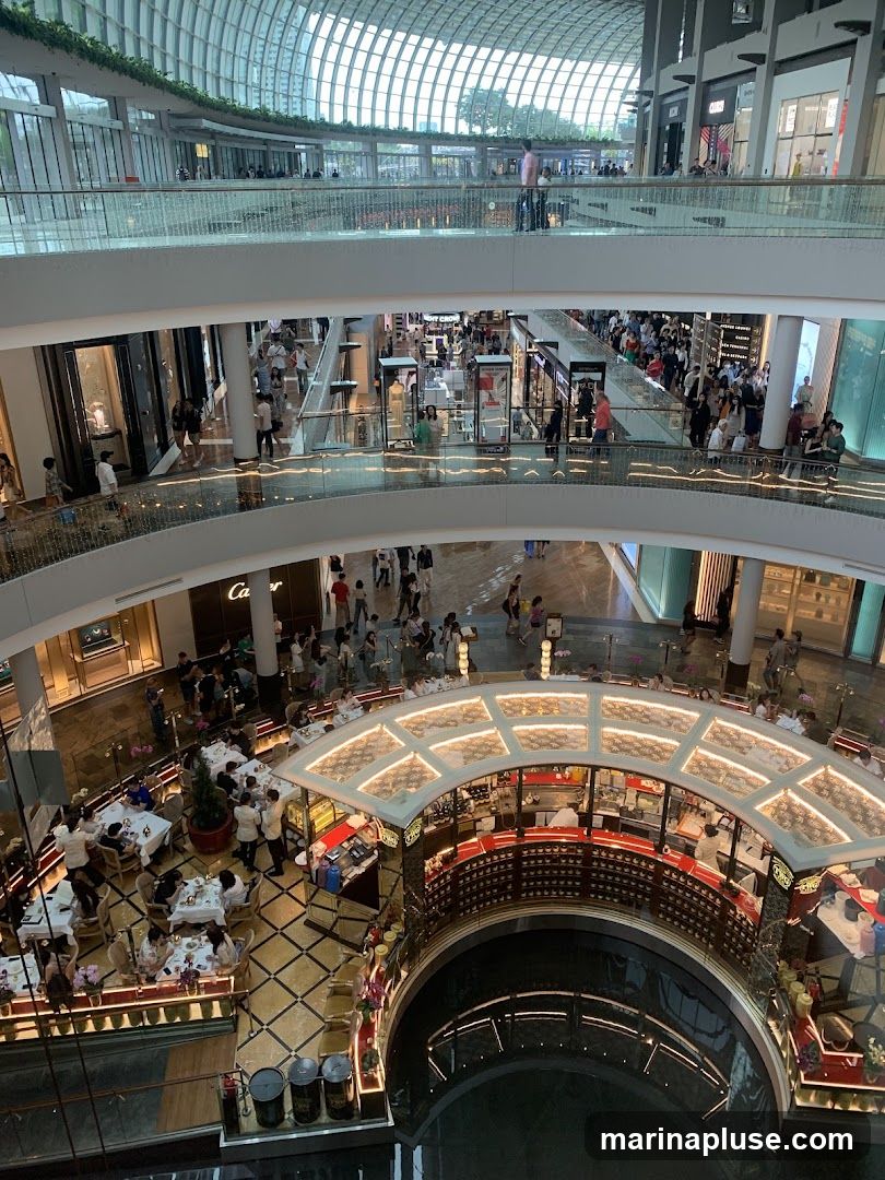 The image shows the interior of a large, modern shopping mall with multiple levels. A large, curved glass roof allows natural light to enter. People are seen walking on the elevated walkways and browsing in the shops. In the foreground, a circular restaurant or cafe area is visible with tables and chairs set up, and a bar area with staff serving customers. The overall impression is one of a busy and upscale retail environment.