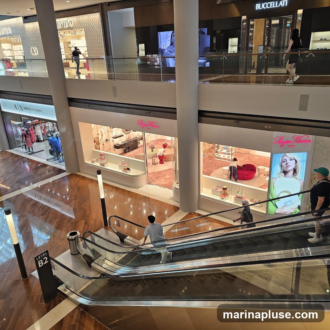 An interior view of a multi-level shopping mall. Escalators are visible in the foreground with shoppers ascending. Several storefronts are displayed on the upper levels, including Valentino, Armani Exchange, and Roger Dubuis. The mall features polished wooden floors and modern architectural elements.