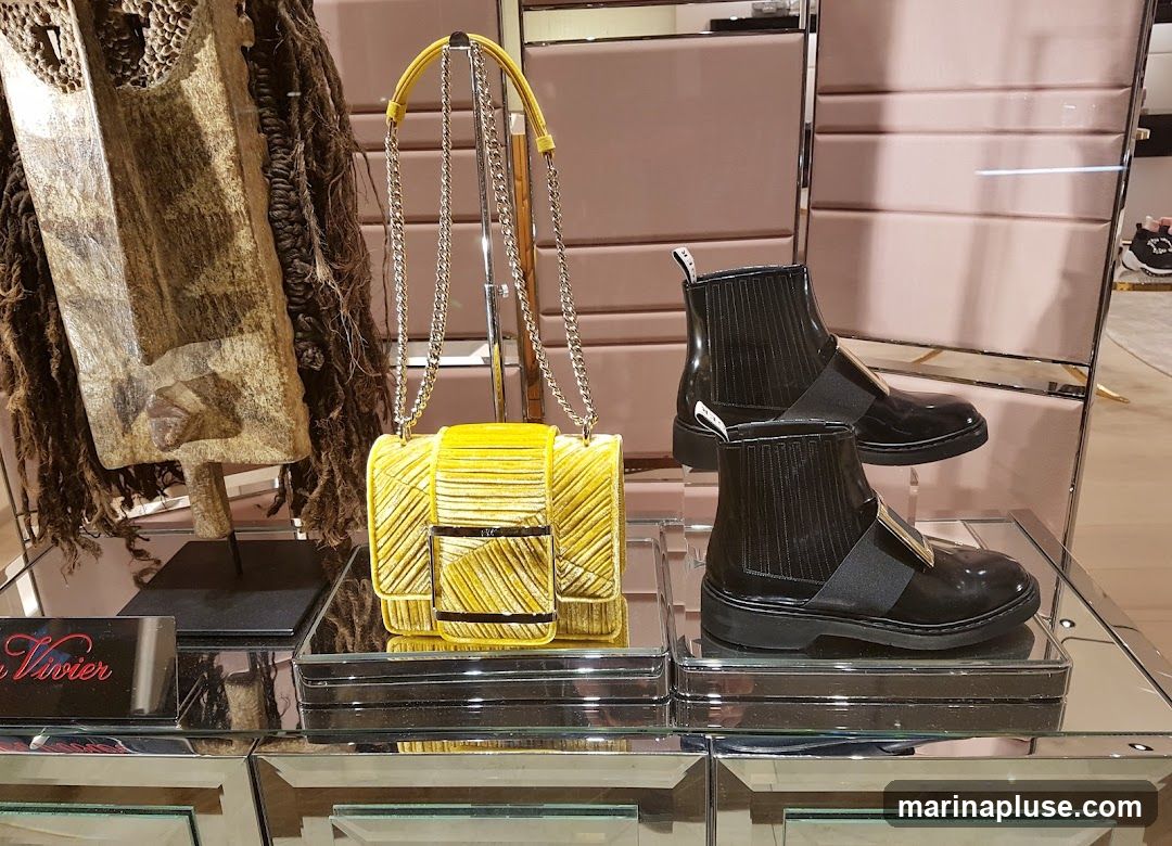 A display case features a yellow velvet handbag with a chain strap and two pairs of black patent leather ankle boots. To the left, a decorative item resembling a mask with dark, textured material is visible. A small black plaque with red script text is in the foreground. The background shows a pink, textured wall and mirrored surfaces.