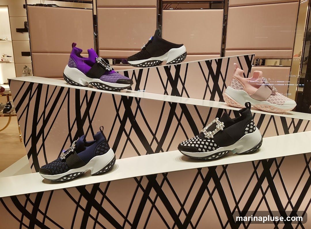 A display of several pairs of women's fashion sneakers on tiered shelves. The sneakers feature decorative buckle embellishments and come in various colors including purple, black, pink, and navy blue. The display has a modern aesthetic with a pink and black geometric pattern on the shelving.