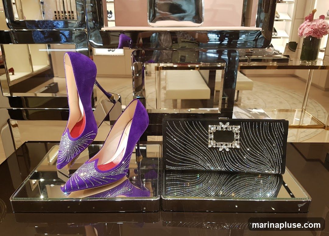 A display of luxury fashion accessories, featuring a pair of vibrant purple suede high-heeled shoes adorned with sparkling embellishments and a black clutch bag with a similar jeweled clasp and sequined pattern. The items are showcased on mirrored surfaces within a well-lit retail environment, with blurred background elements suggesting a high-end boutique.