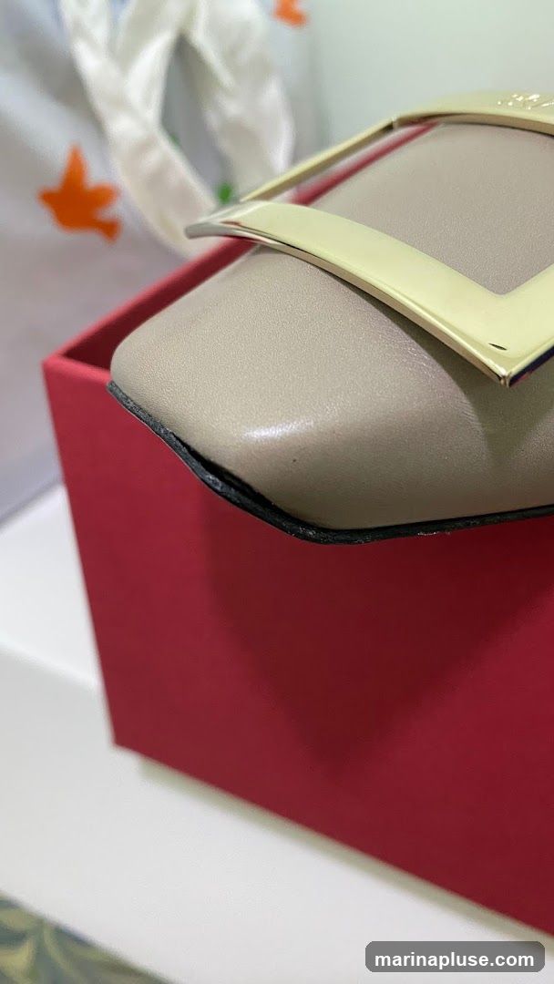 A close-up shot of a beige shoe with a prominent gold buckle. The shoe is resting on a red box. In the background, a white cloth with orange bird-like figures is visible.