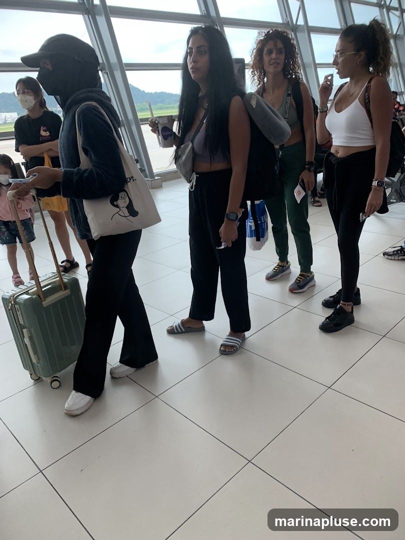 A group of people, mostly women, are standing in line inside an airport terminal. One person is pulling a suitcase, and others are holding drinks or bags. The architecture of the terminal is visible with large windows and a modern design.