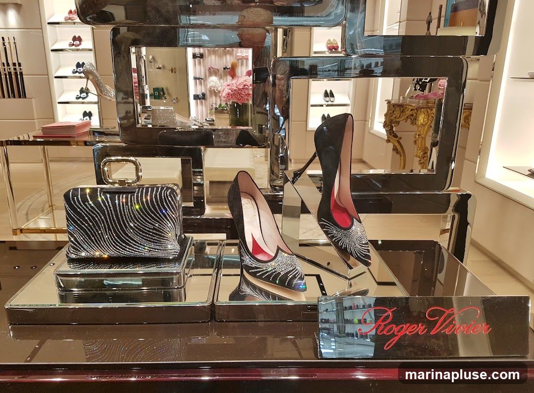 A display of luxury footwear and handbags from Roger Vivier. In the foreground, a black clutch adorned with sparkling crystals is showcased on a mirrored platform. To its right, a pair of black high-heeled pumps, also embellished with crystals, are displayed. The background reveals shelves stocked with more shoes and accessories, along with decorative floral arrangements. A prominent red Roger Vivier logo is visible on a reflective surface in the lower right corner.