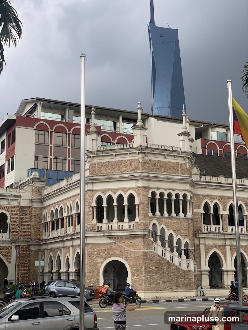 A view of a historic, ornate brick building with arched windows and a grand staircase, set against a cloudy sky. In the background, a modern, blue glass skyscraper with a distinctive angular design rises prominently. Several vehicles, including cars and motorcycles, are visible on the street in front of the historic building.