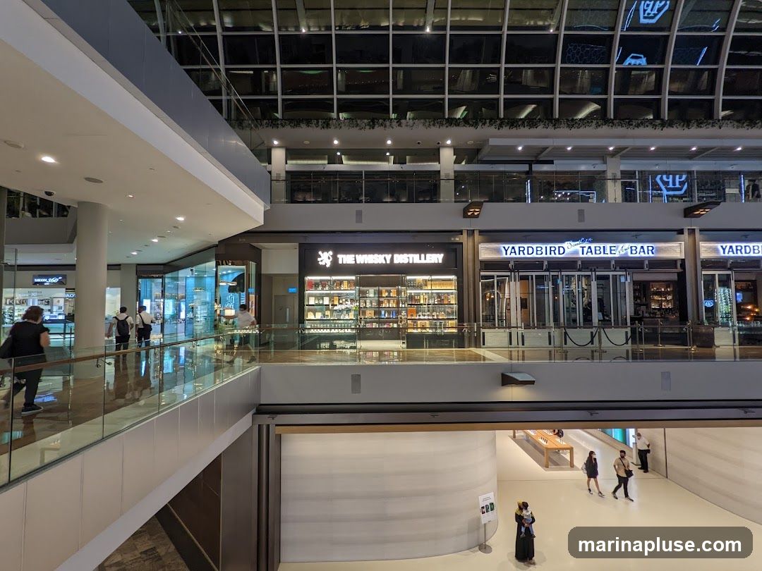 The image shows the interior of a multi-level shopping mall. On the upper level, several storefronts are visible, including 'THE WHISKY DISTILLERY' and 'YARDBIRD TABLE & BAR'. The architecture features a modern design with glass railings and a reflective ceiling. People are seen walking on the different levels. Below, an Apple Store is visible with customers browsing products.