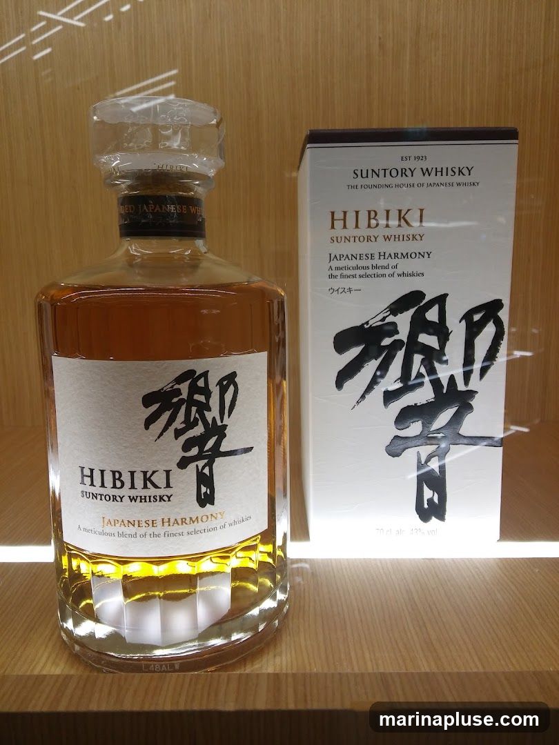 A bottle of Hibiki Japanese Harmony whisky stands next to its matching box on a wooden shelf. The bottle is clear glass with a faceted base, filled with amber-colored whisky. The label on the bottle features Japanese calligraphy and the text "HIBIKI SUNTORY WHISKY JAPANESE HARMONY". The box is white with similar branding and calligraphy.