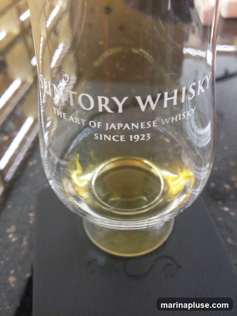A close-up shot of a clear glass containing amber-colored liquid, likely whisky. The glass has white lettering that reads "SUNTORY WHISKY THE ART OF JAPANESE WHISKY SINCE 1923". The glass is resting on a dark surface.