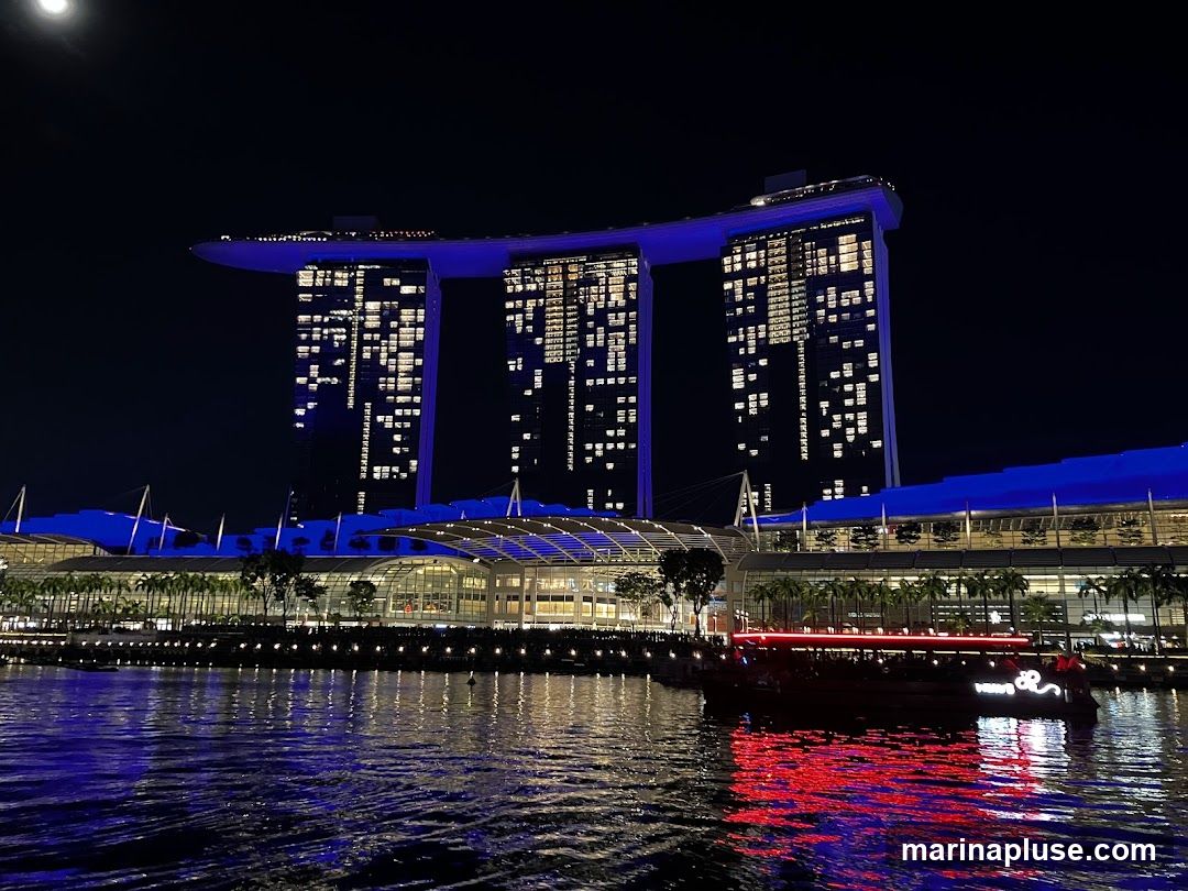 A nighttime view of the Marina Bay Sands complex in Singapore, illuminated with blue lights. The three towers are visible, with many windows lit up. In the foreground, a body of water reflects the lights of the buildings and a boat with red neon lights. The moon is visible in the upper left corner.