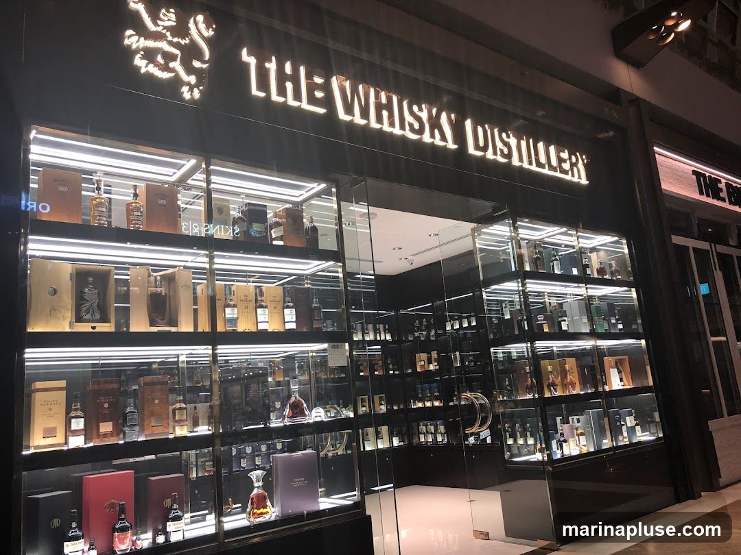 The exterior of a well-lit shop called "The Whisky Distillery" is shown. The storefront features large glass windows displaying numerous bottles of whisky on illuminated shelves. The shop's name is prominently displayed in large, illuminated letters above the entrance.