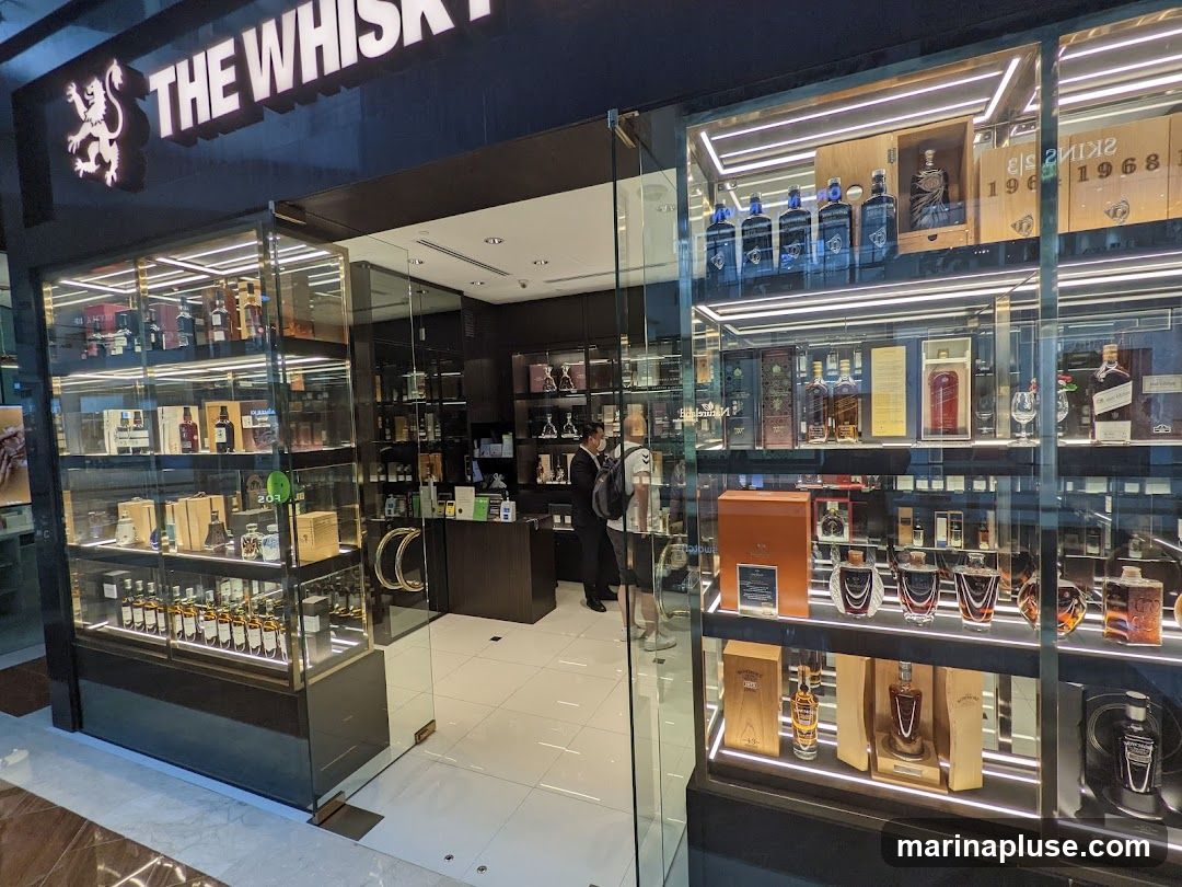 The exterior of a well-lit liquor store specializing in whisky. The store features large glass display windows showcasing numerous bottles of whisky arranged on shelves. The store's name, "THE WHISKY", is prominently displayed in large illuminated letters above the entrance. A person can be seen inside the store near the counter.