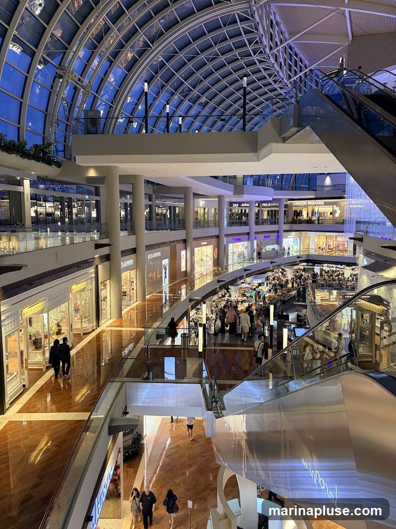 The image shows the interior of a large, modern shopping mall with multiple levels. A distinctive glass and metal arched ceiling covers the space, allowing natural light to filter in. Escalators and walkways connect the different floors, which are lined with various retail stores. People are seen browsing and walking through the mall. The polished wooden floors reflect the ambient lighting, creating a warm and inviting atmosphere.
