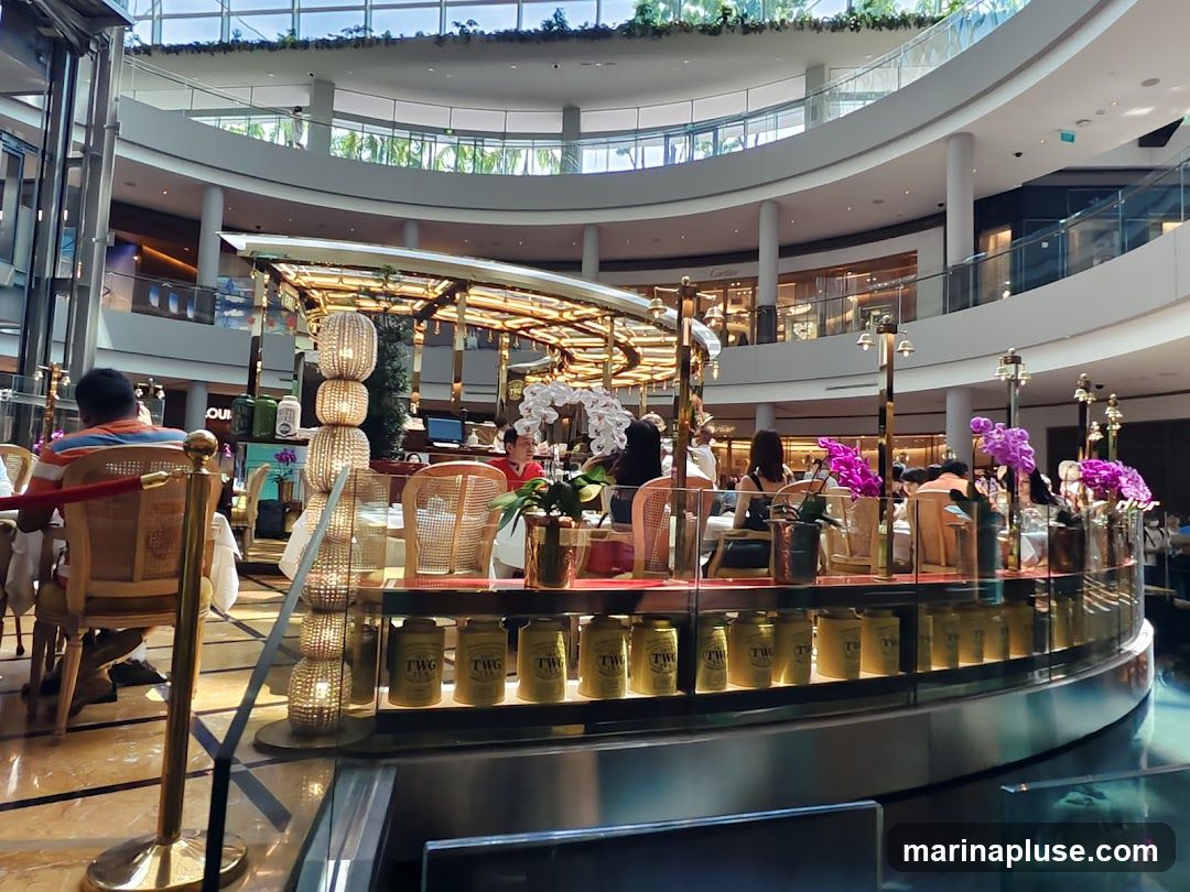 An indoor cafe setting within a large, multi-level shopping mall. Several patrons are seated at tables, some of which are adorned with floral arrangements. The cafe features decorative lighting and displays of tea canisters. In the background, other shops and escalators are visible, indicating a bustling commercial environment.