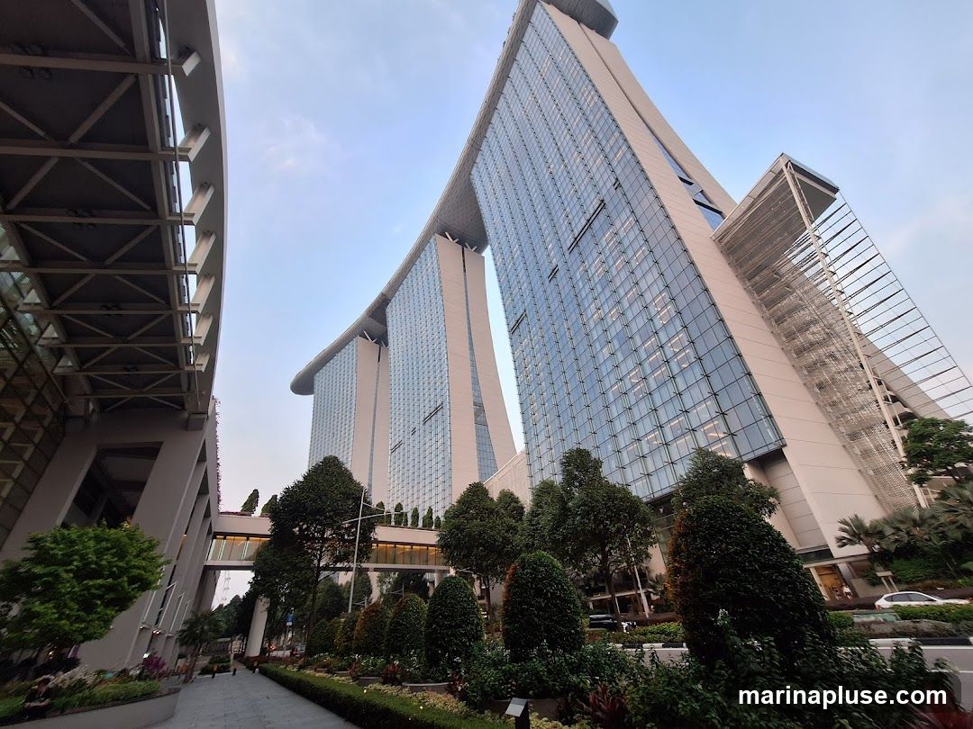 A low-angle shot of the Marina Bay Sands hotel in Singapore, with its distinctive three towers connected by a sky-high platform. The glass facade of the buildings reflects the sky. In the foreground, there are manicured gardens and trees, and a walkway. The sky is clear and pale blue.