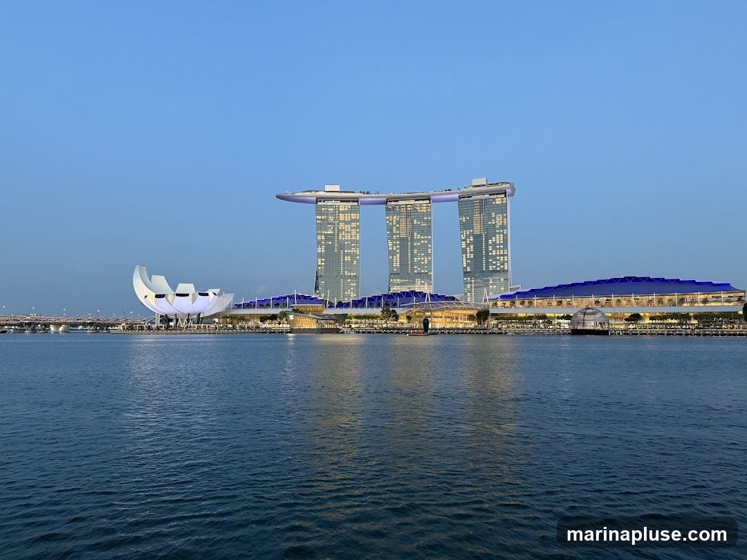 A wide shot of the Marina Bay Sands complex in Singapore, viewed across the water at dusk. The iconic three towers, connected by the SkyPark, are illuminated, as are the surrounding buildings and the ArtScience Museum. The sky is a clear, deep blue, and the water reflects the lights of the city.
