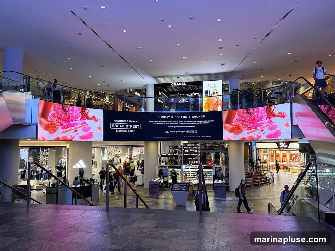 A wide shot of a modern shopping mall interior. A large digital screen displays an advertisement for "Gordon Ramsay Bread Street Kitchen & Bar" promoting "Sunday Kids' Fun & Brunch." Above this, signs for "Chanel" and "N°5" are visible. Below the main screen, storefronts for Adidas and other retailers can be seen. People are walking on escalators and through the mall.
