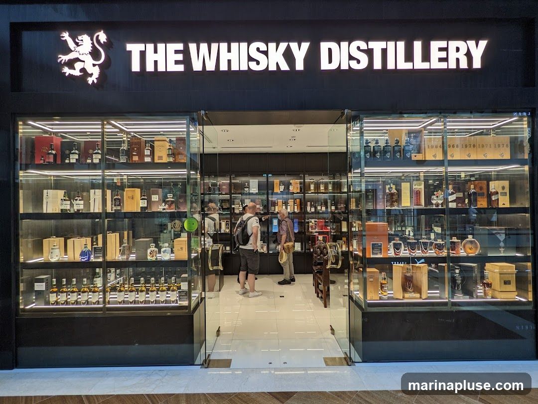 The exterior of a liquor store called The Whisky Distillery. The store has a large glass storefront displaying numerous bottles of whisky on shelves. Two men are standing inside the store, looking at the bottles. The store's name and logo are visible above the entrance.