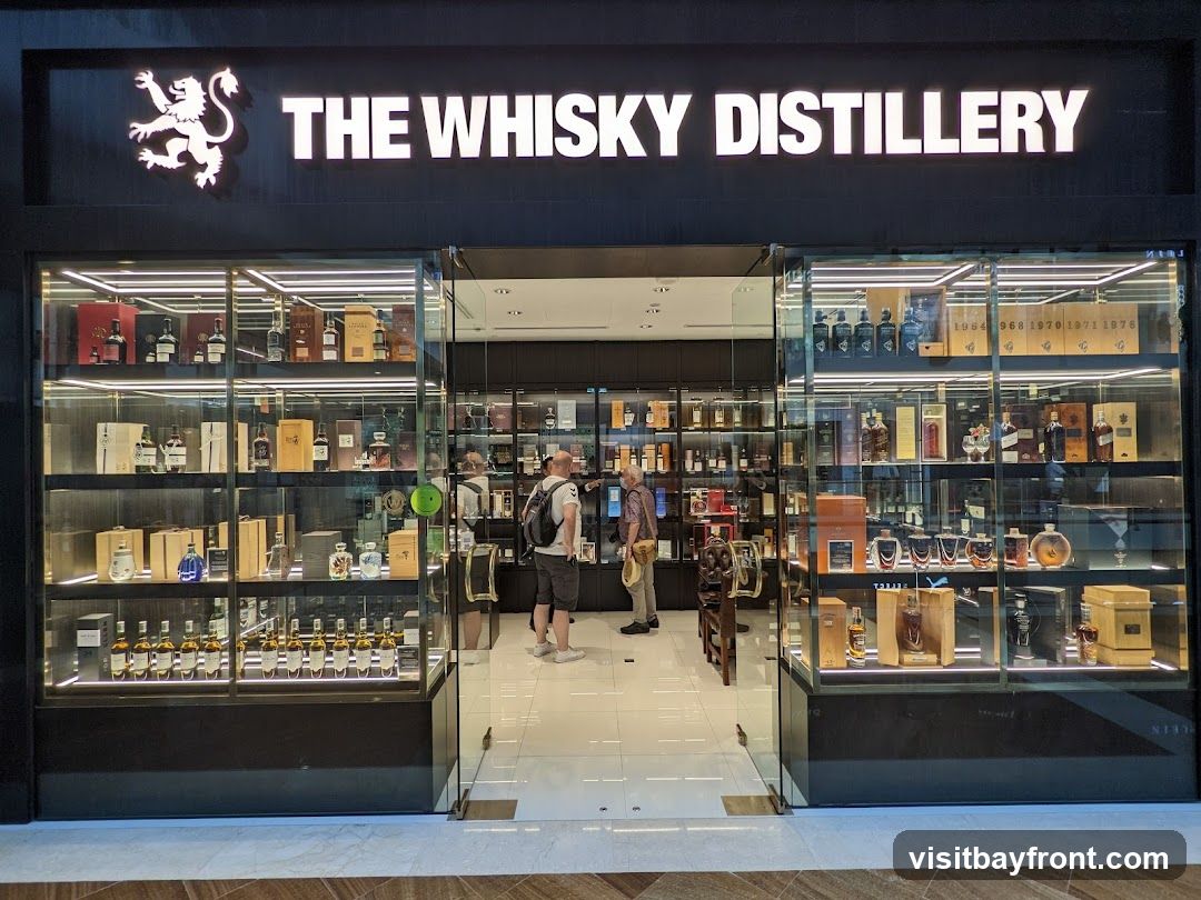 The exterior of a liquor store called The Whisky Distillery. The store has a large glass storefront displaying numerous bottles of whisky on shelves. Two men are standing inside the store, looking at the bottles. The store's name and logo are visible above the entrance.