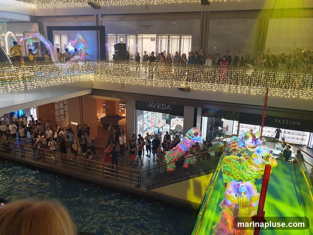 A vibrant indoor scene with a performance featuring colorful dragon costumes, likely a cultural celebration. Spectators are gathered on multiple levels, watching the event. The architecture suggests a modern shopping mall with decorative lighting. Stores like AVEDA and PAZZION are visible in the background.