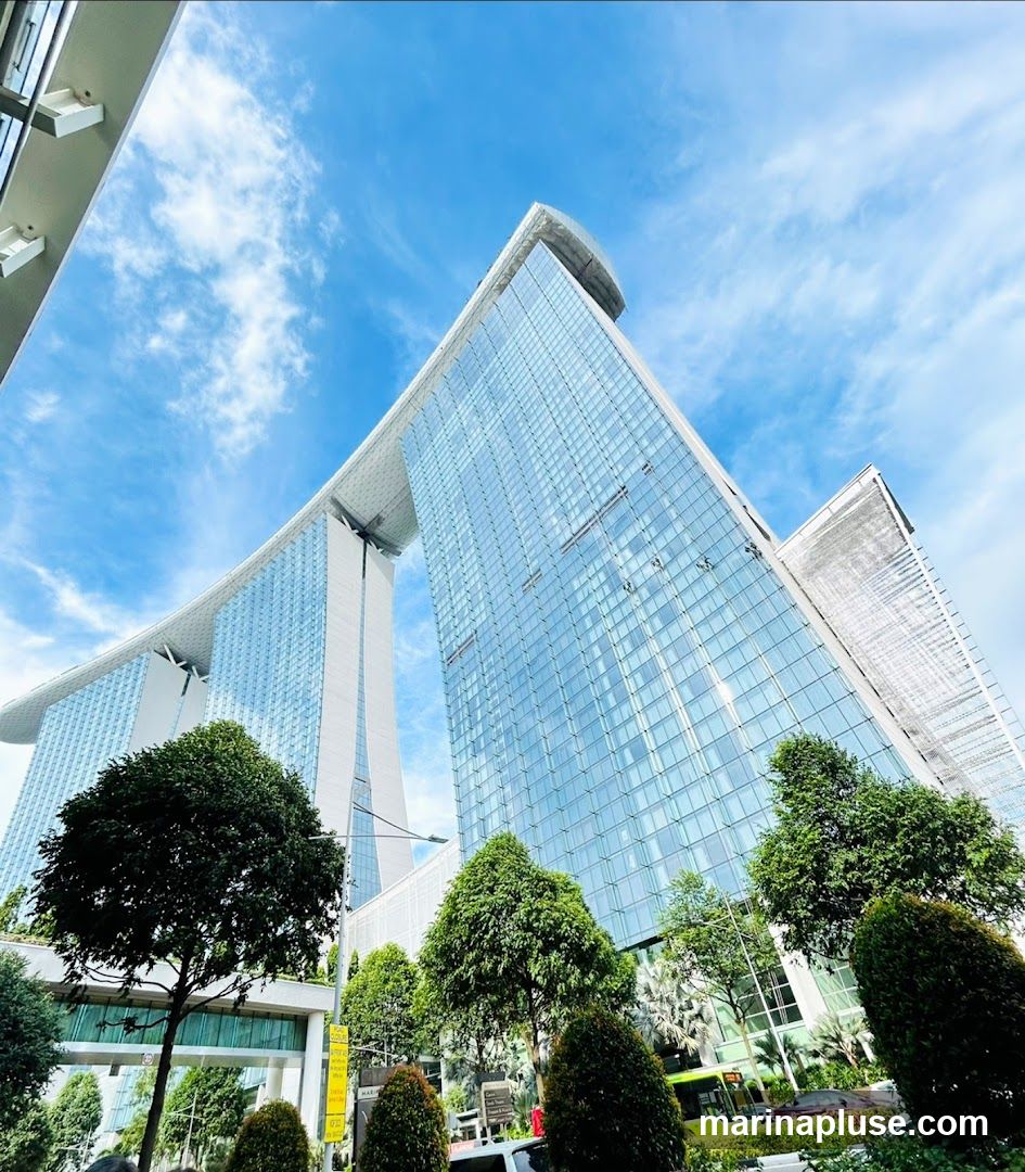 A low-angle shot of the iconic Marina Bay Sands hotel in Singapore, with its distinctive three towers connected by a sky-high platform. The glass facade of the buildings reflects the bright blue sky and scattered white clouds. Lush green trees and manicured bushes are visible in the foreground, with a street and some vehicles in the background.