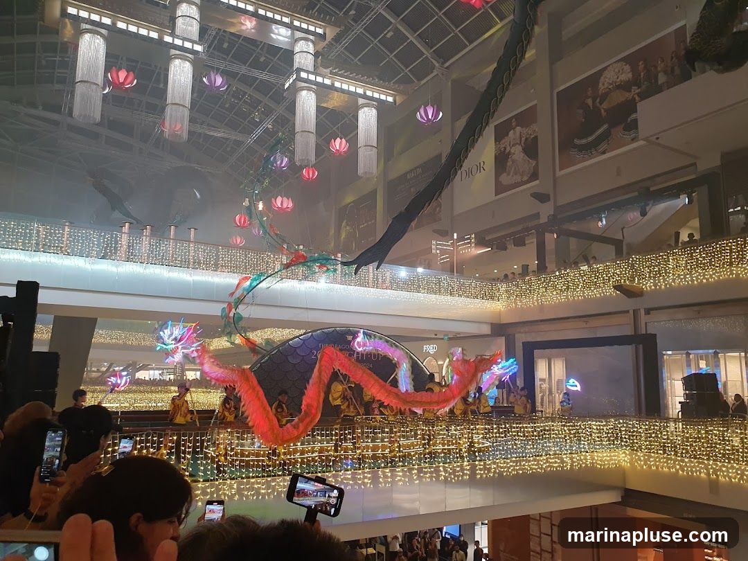 A vibrant dragon dance performance is taking place in a multi-level shopping mall. The dragon, made of colorful fabric, is being manipulated by performers dressed in yellow. The mall is decorated with numerous strings of warm white fairy lights along the railings of each level. Overhead, large, modern chandeliers hang from the ceiling, and colorful lotus-shaped lanterns are suspended in the air. Large advertisements, including one for Dior, are visible on the upper levels. Spectators are gathered on the lower levels, many holding up their phones to capture the event.