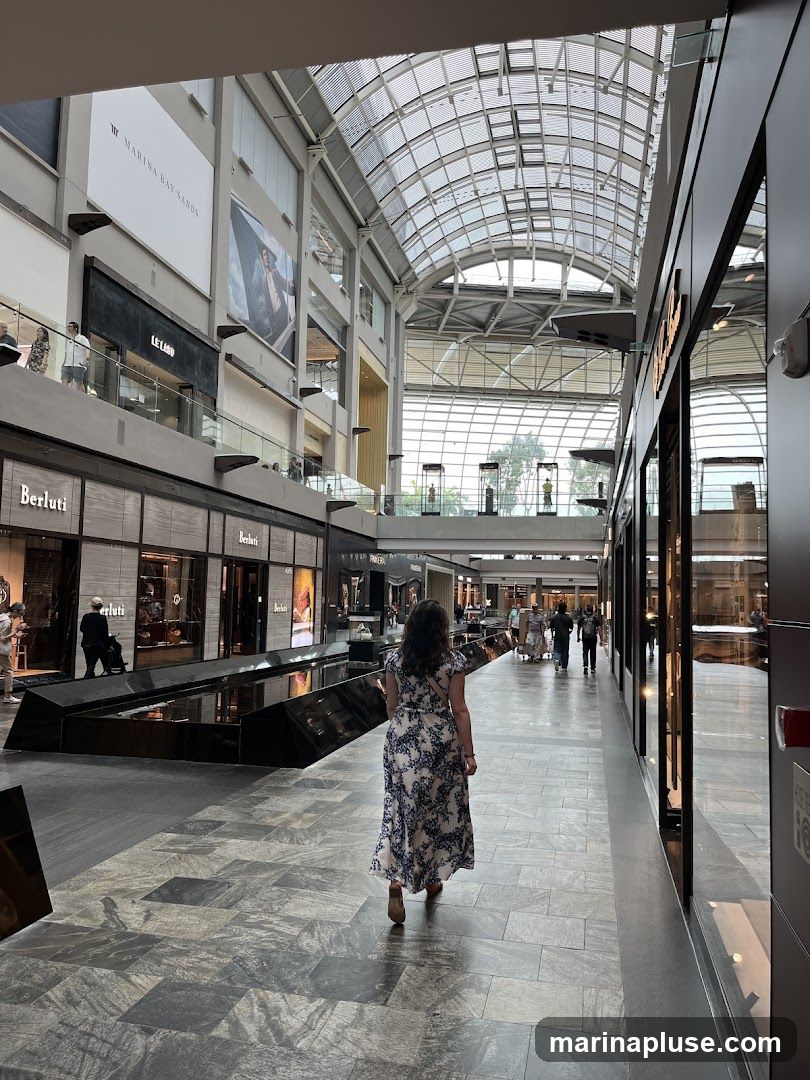 A woman in a floral dress walks down a wide, well-lit corridor in a modern shopping mall. The mall features a high, arched glass ceiling, allowing natural light to flood the space. On the left, several luxury storefronts are visible, including "Berluti" and "Le Labo." Large advertisements and signage are displayed on the upper levels, including one for "Marina Bay Sands." On the right, more storefronts with reflective facades line the walkway. Other shoppers can be seen in the distance.