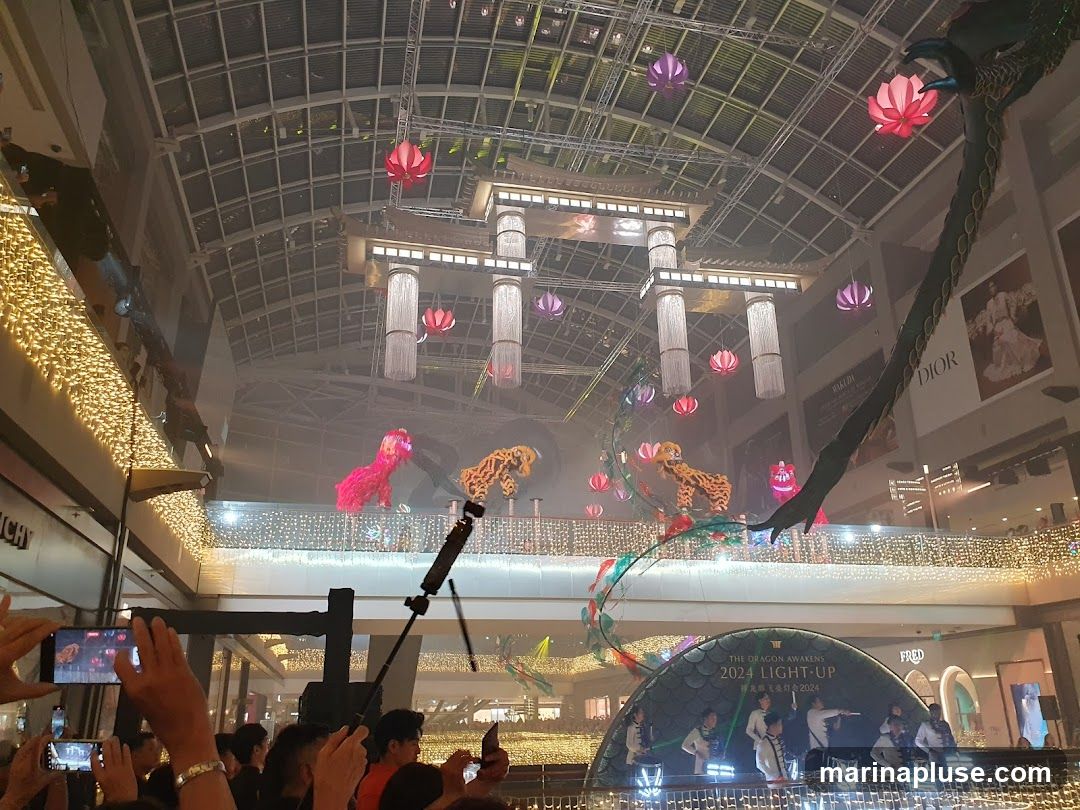 A festive indoor scene with elaborate decorations for Lunar New Year. Lion dancers in vibrant costumes perform on a raised platform, while a large dragon sculpture hangs from the ceiling. The space is adorned with numerous lotus-shaped lanterns and twinkling string lights. A crowd of people are visible in the foreground, many holding up their phones to capture the event. Signage for brands like Dior and Fred is visible in the background, suggesting this is a shopping mall.