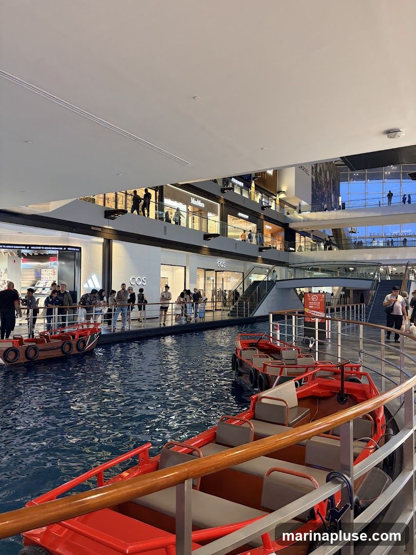 An indoor shopping mall features a canal with red boats for rides. Shops with visible signage like COS, Max Mara, and Chanel are on multiple levels. People are seen browsing and walking around the mall.